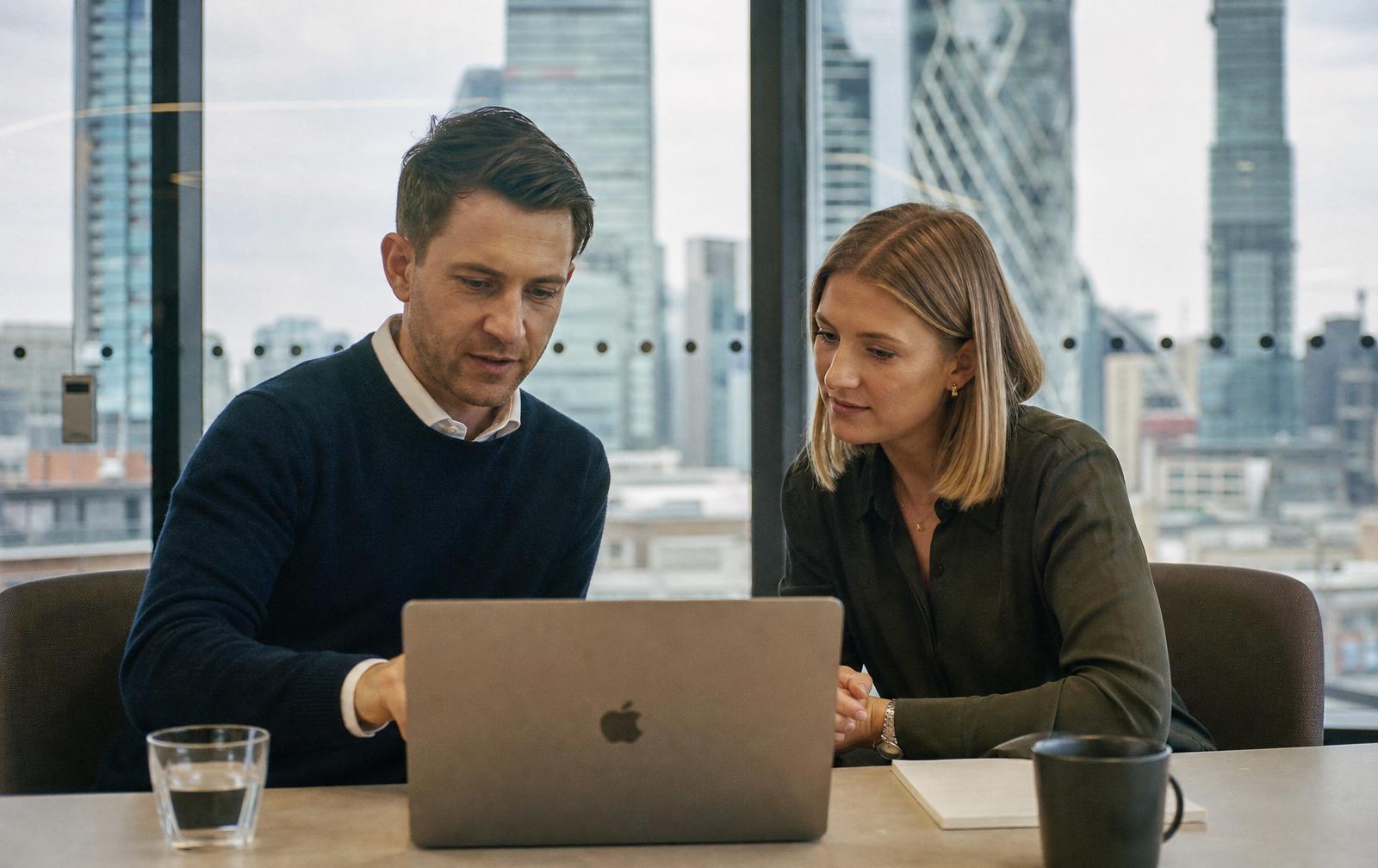 Two UK business professionals collaborating in a modern London office meeting room, reviewing finance data on a laptop